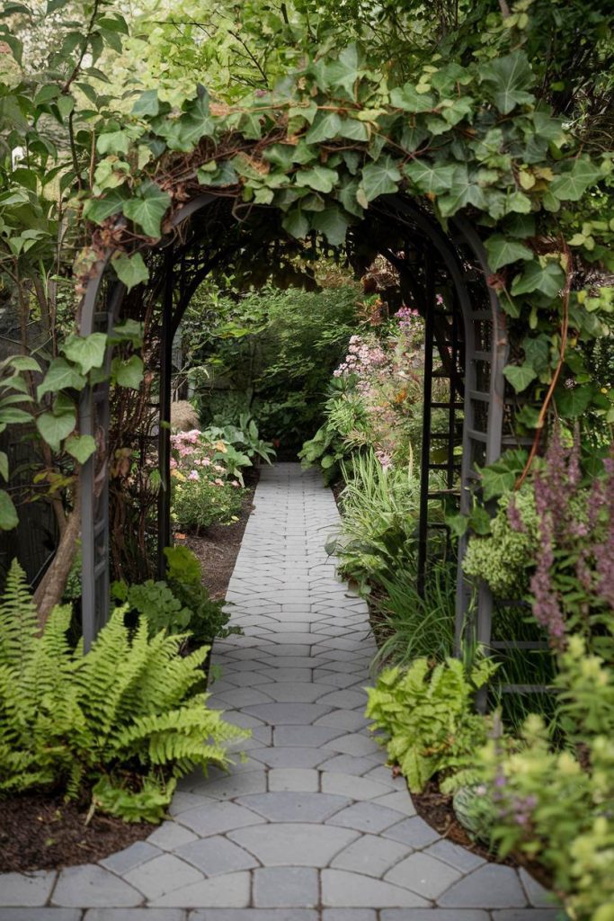 A stone path leads through a garden archway covered in lush greenery, surrounded by various plants and flowers.