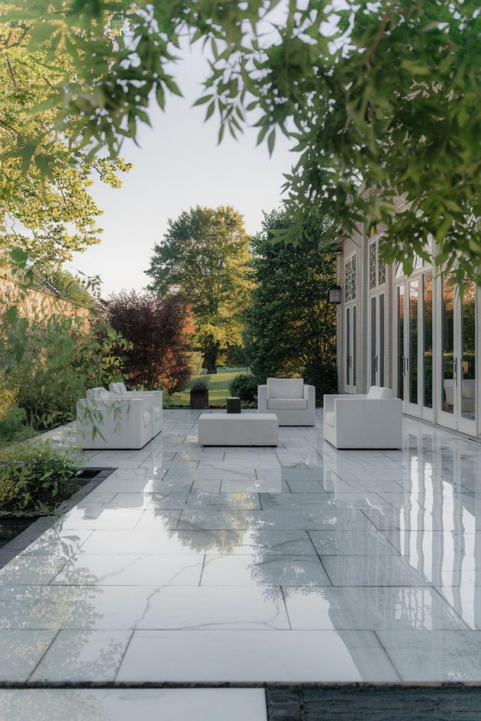 A modern terrace with white furniture on a polished stone floor surrounded by lush greenery and trees.