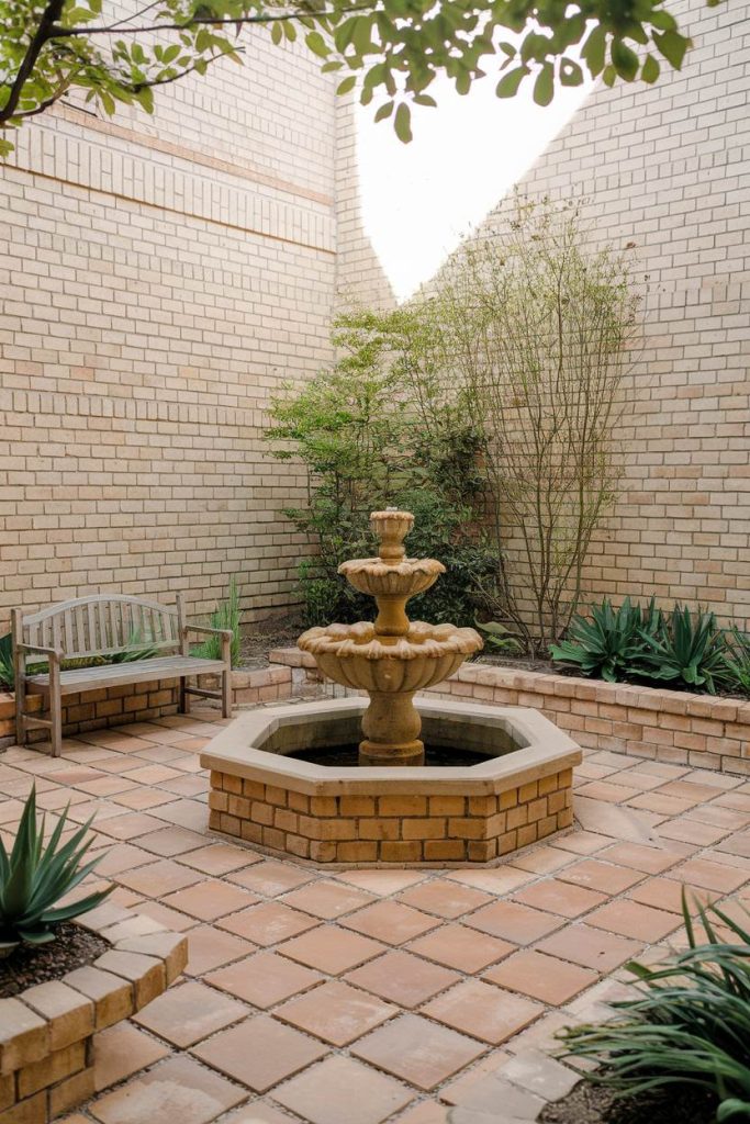 A small courtyard features a central stone fountain surrounded by potted plants, a wooden bench and brick walls.