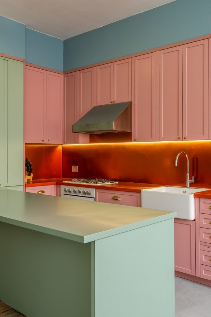 A kitchen with pastel pink cabinets, copper backsplash and a green island. It includes a farmhouse sink, cooktop with extractor hood and under cabinet lighting.