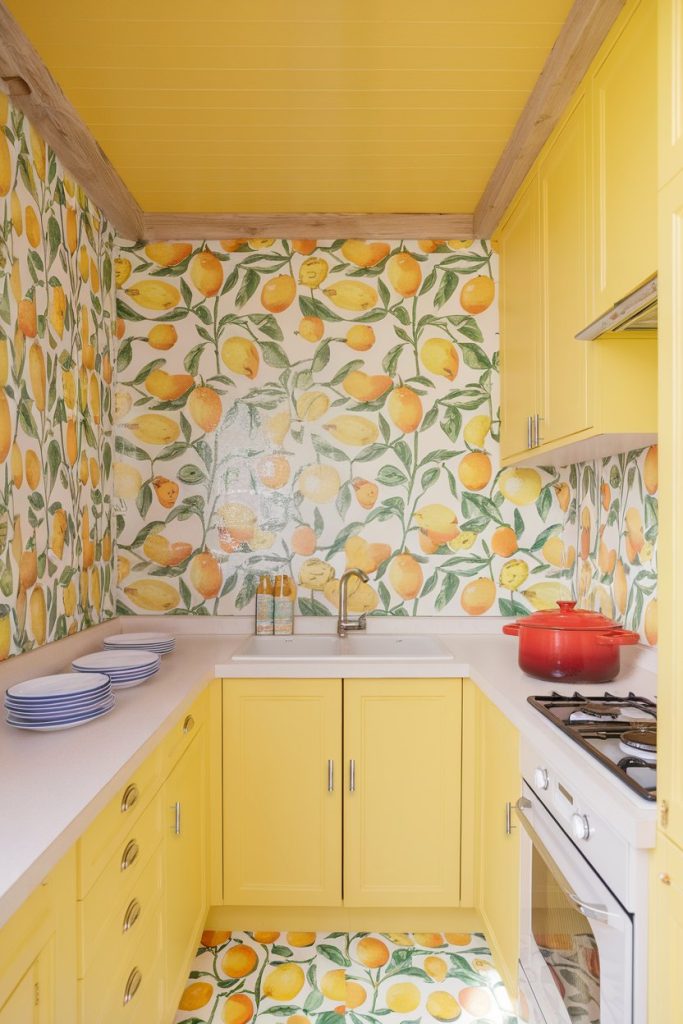 A small kitchen with yellow cabinets and citrus-themed tiles. It has a white countertop, a red pot on the stove and a stack of blue plates.