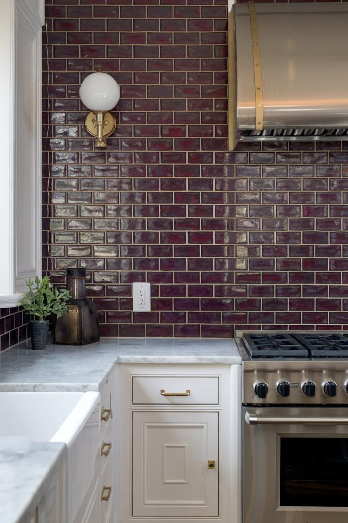 Kitchen with glossy dark red subway tile backsplash, stainless steel stove with hood, white cabinets and marble countertops. Wall lamp and small plant on the counter.