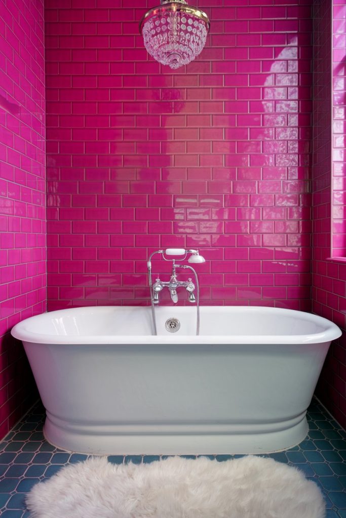 A freestanding white bathtub sits in a bathroom with bright pink tiled walls and a chandelier above. There is a fluffy white carpet on the blue tiled floor.