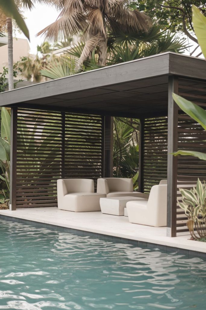 Poolside cabana with modern furniture surrounded by tropical plants. The structure features wooden slatted walls and a flat roof that provides a shaded seating area next to the pool.