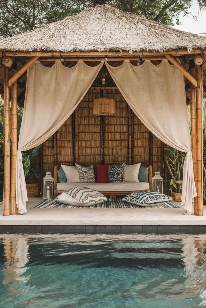 A tropical cabana with a bamboo structure, white curtains and blue patterned cushions overlooks a pool surrounded by greenery.