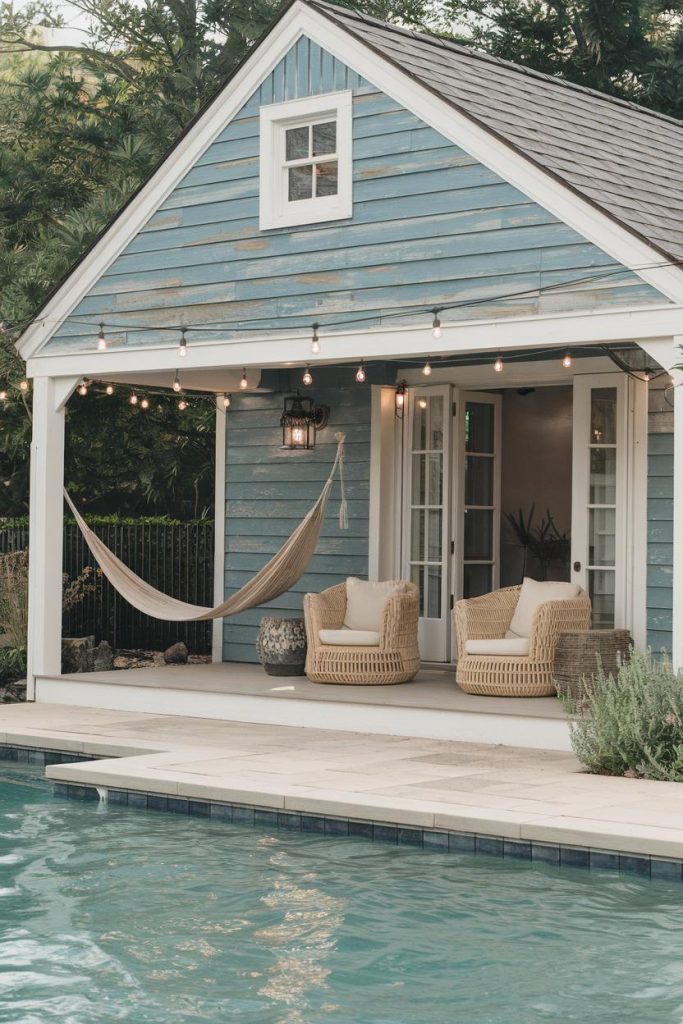 A cozy blue poolside cabana with wicker chairs, a hammock, fairy lights and a clear swimming pool in the foreground.