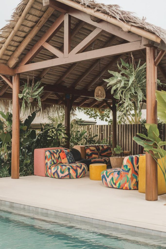 A tropical-style outdoor lounge with colorful chairs under a thatched gazebo surrounded by lush greenery near a pool.