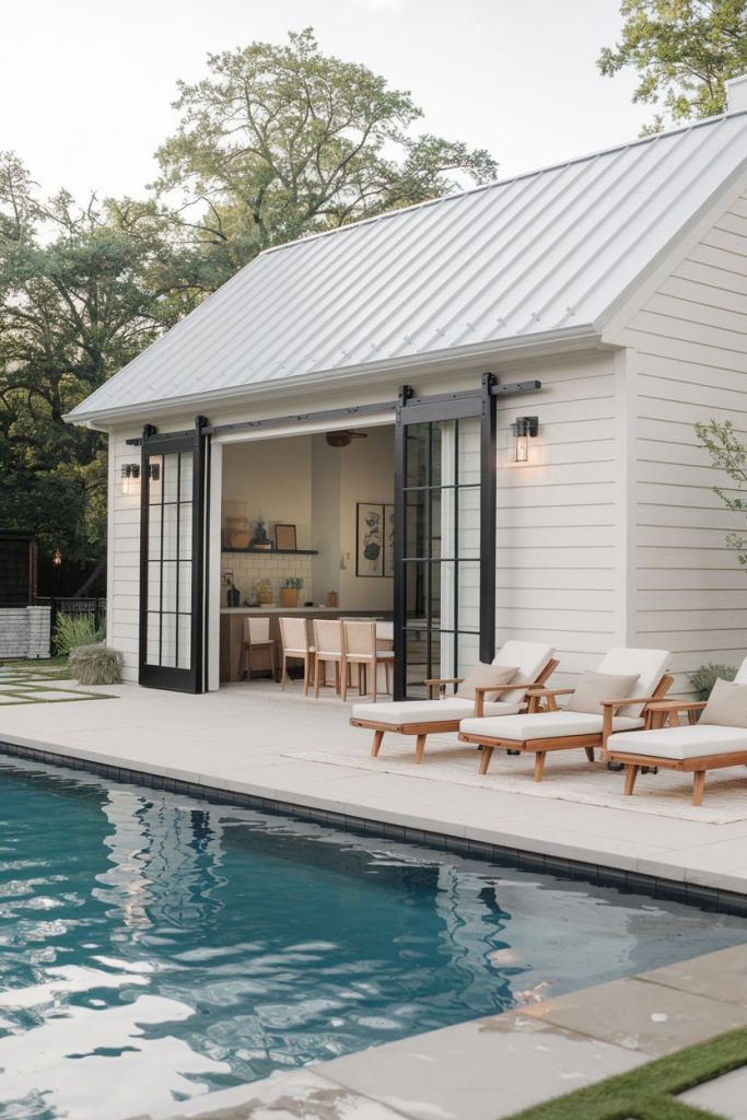 A modern house with large glass doors leading to a poolside terrace. There are three wooden lounge chairs with cushions next to the pool. Trees can be seen in the background.