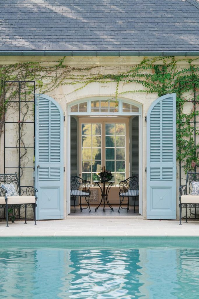 Open French doors with light blue shutters lead to a cozy seating area. Wrought iron chairs and a table overlook a tranquil swimming pool. Lush vines grow along the stone outer walls.