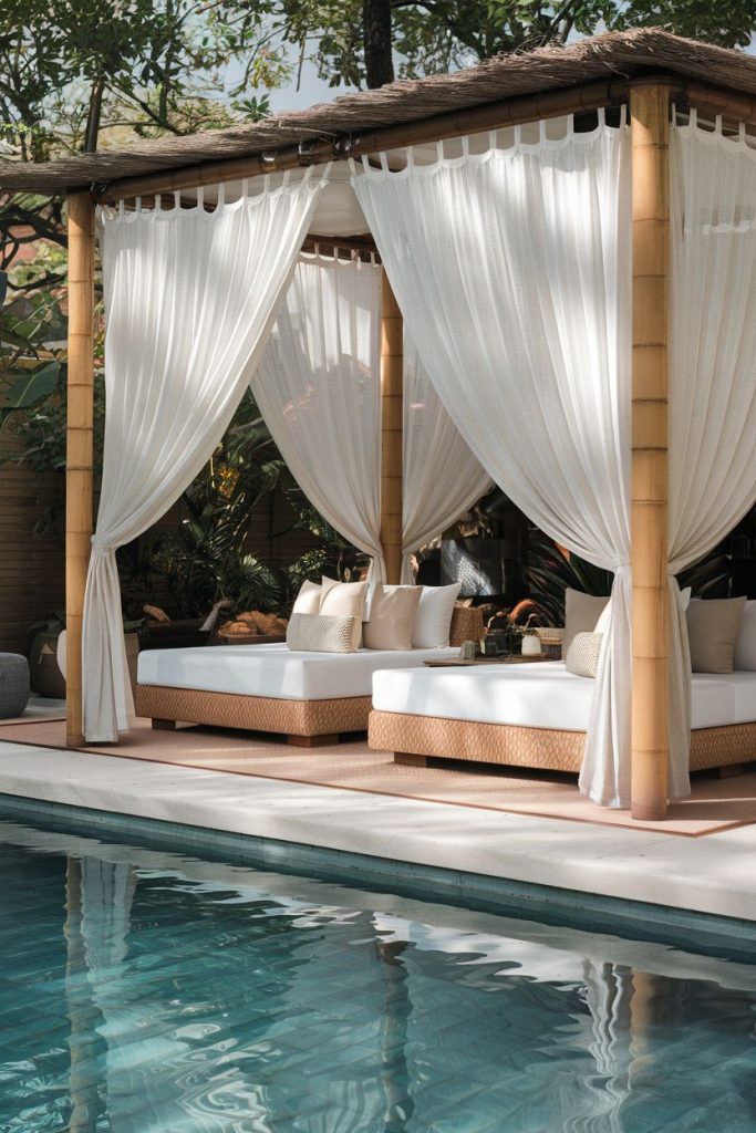 A poolside cabana with white curtains and cushioned seating sits next to a clear blue pool surrounded by greenery.