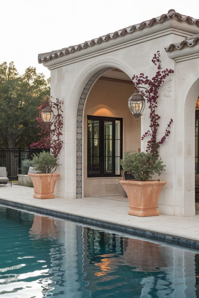 Elegant poolside stone pavilion with arched openings, lanterns and potted plants. Tile accents decorate the entrance and the pool reflects the structure. Trees are in the background.