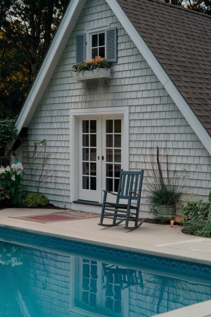 A rustic house with a shingled roof lies by a pool. A rocking chair sits on the poolside patio and a flower box adorns the window above the French doors.