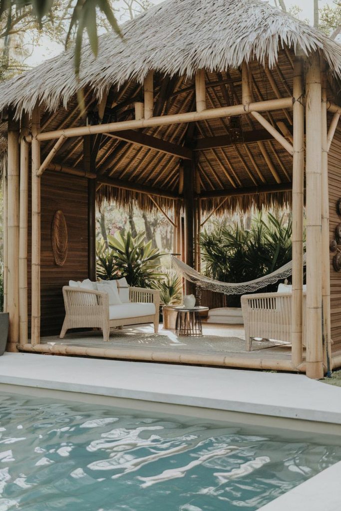 A bamboo pavilion with a thatched roof offers outdoor seating and a hammock. It is surrounded by tropical plants and overlooks a swimming pool.