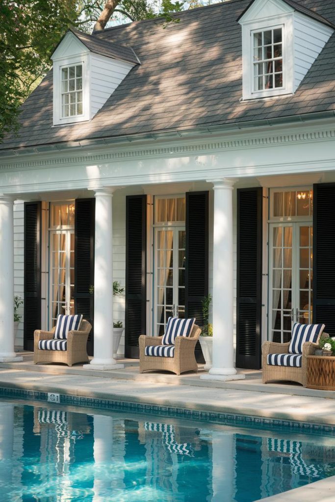 A Colonial-style home with white columns and black shutters features a pool in the foreground and patio chairs with striped cushions.