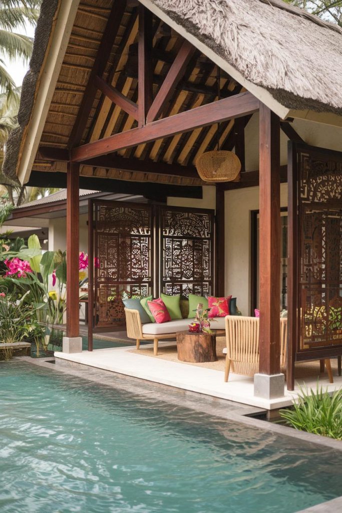 A tropical outdoor poolside terrace with a thatched roof, wooden furniture, colorful cushions and intricate wooden panels amidst lush vegetation.