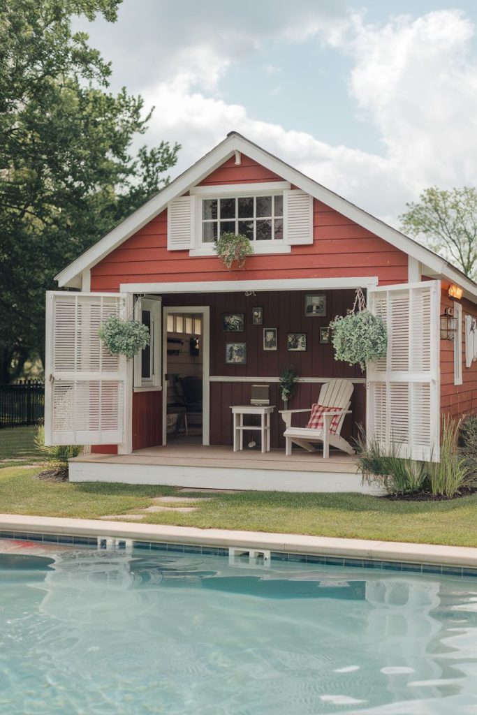 A red pool house with open white shutters surrounded by greenery stands next to a pool. Chairs and a small table are on the porch. The sky is partly cloudy.