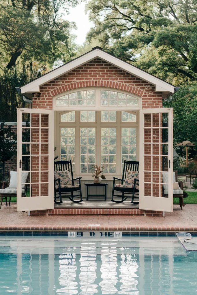 A small brick pavilion with open glass doors features two rocking chairs and a table overlooking a swimming pool surrounded by trees.