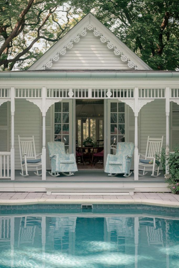 A white Victorian-style porch with rocking chairs overlooks a swimming pool surrounded by greenery and trees.