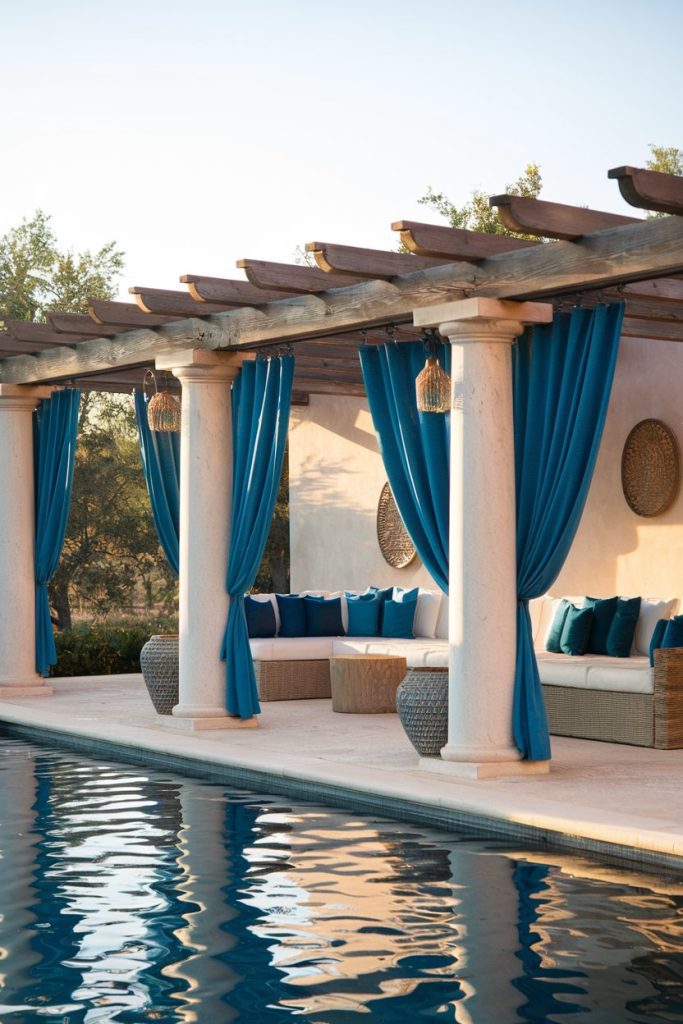 A poolside seating area with white columns and blue curtains. Wicker furniture with blue and white cushions is arranged under a wooden pergola. The pool reflects the scene.