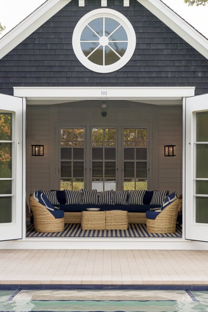 Poolside open plan room with wicker furniture, blue and white striped cushions and round window above. French doors lead to the seating area overlooking a tranquil pool.