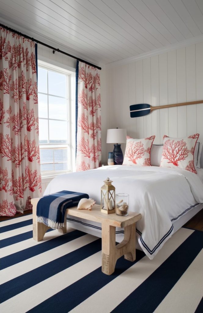 A bright bedroom with nautical decor, a striped rug, coral-print curtains and pillows, a hanging rudder and ocean views through a large window.