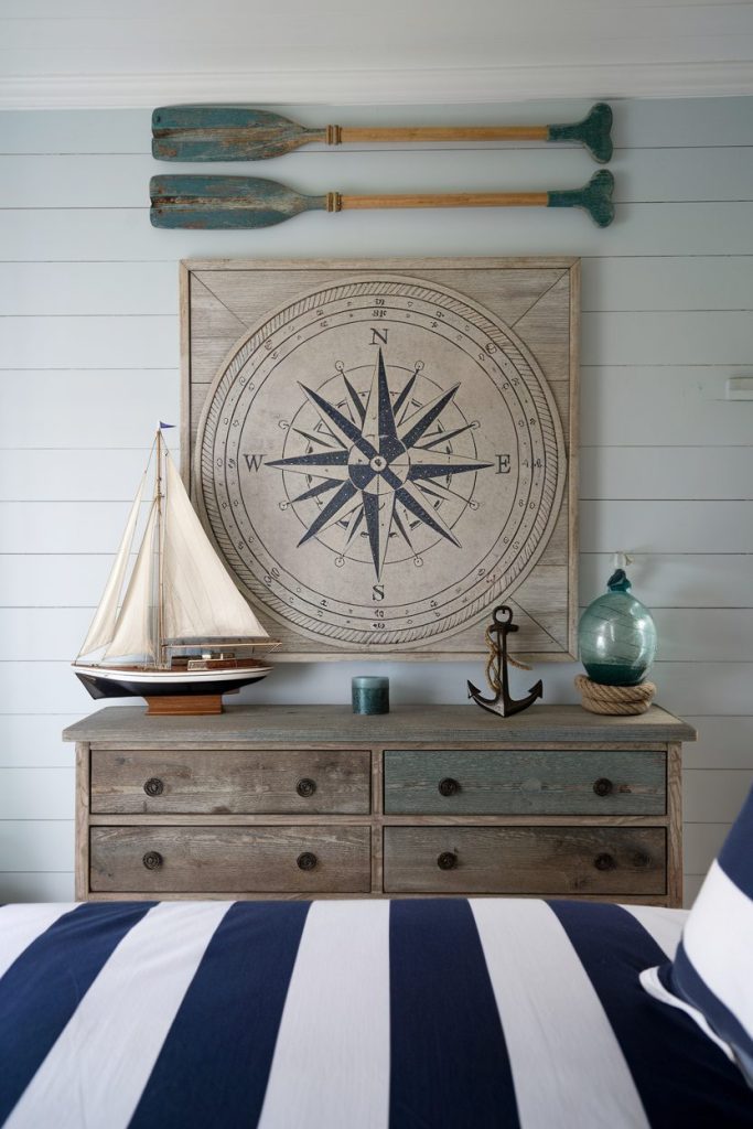 Nautical-themed bedroom with compass wall art, wooden oars, sailboat model, anchor and blue striped bedding on a wooden dresser.