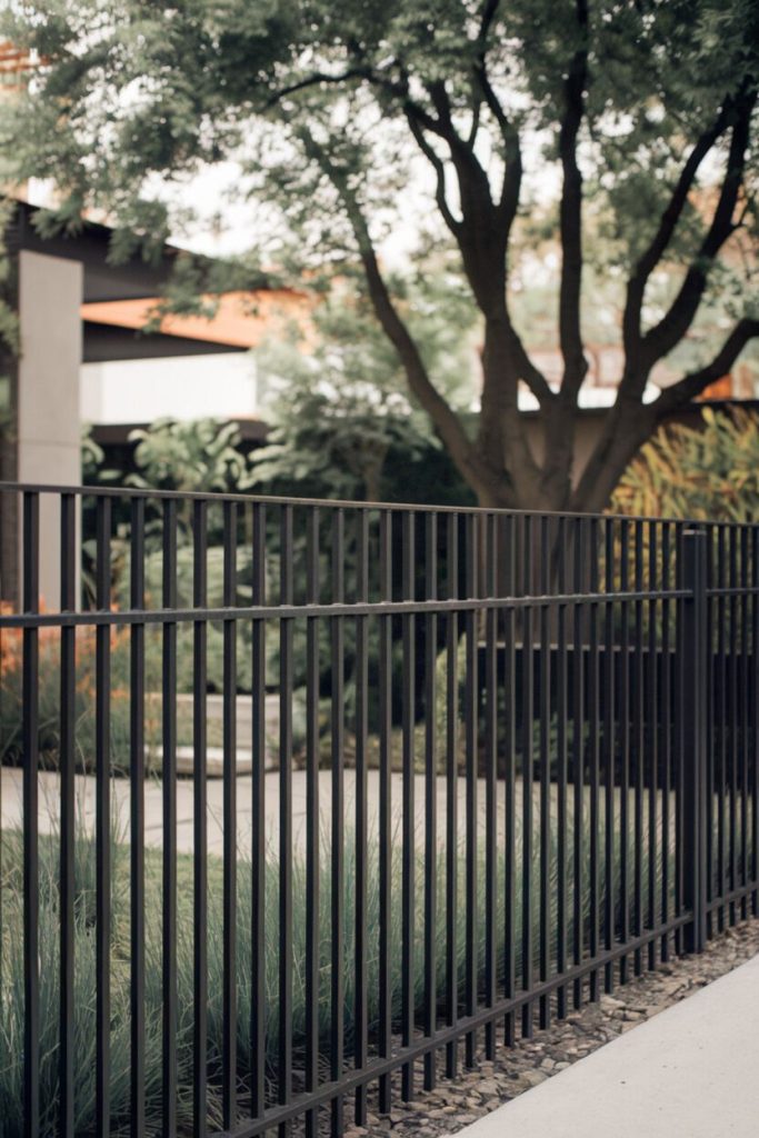 Black metal fence in front of a landscaped garden with grass and a large tree. Buildings can be seen in the background.