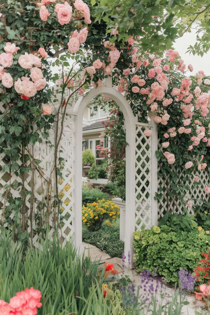 A garden with a white lattice archway decorated with pink roses leading to a path surrounded by various colorful flowers and greenery.