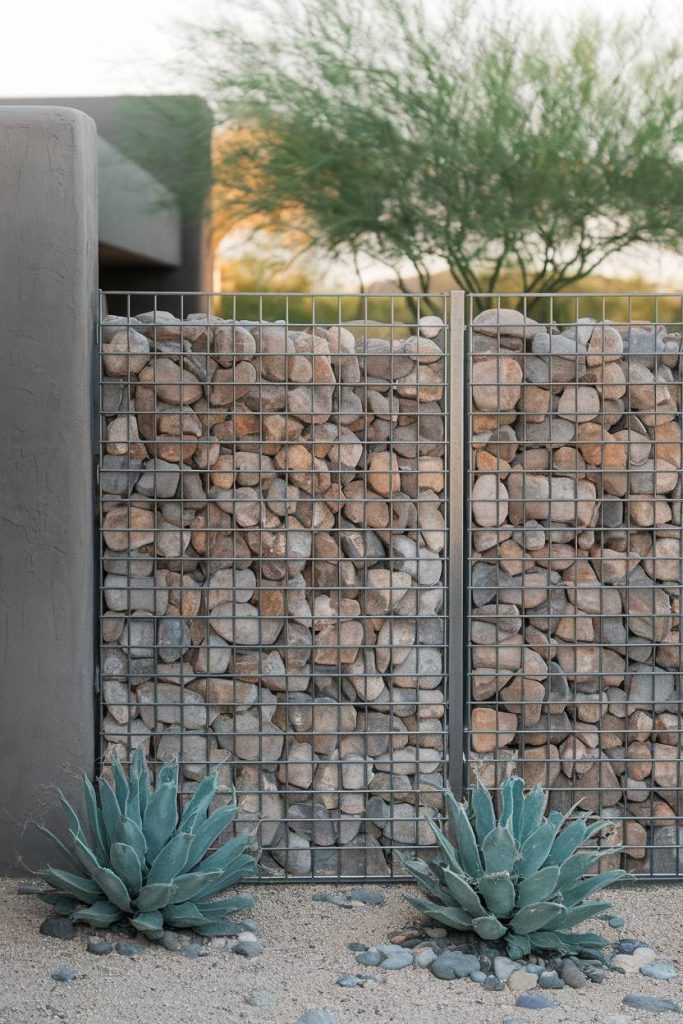 A stone-filled gabion wall lies in a desert landscape, flanked by two succulents, with a blurred tree in the background.