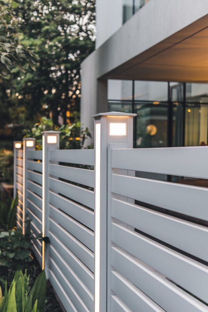 Modern white fence with illuminated posts in front of a building with large windows and green spaces.