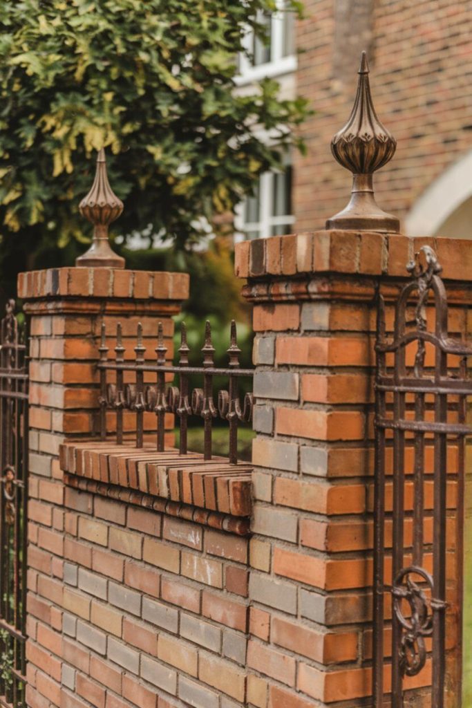 A brick fence with ornamental ironwork and pointed finials against a blurred background of a building and trees.