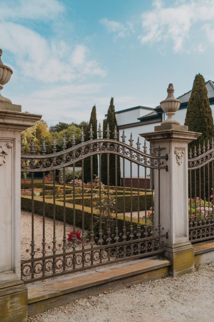 Ornate metal gate in front of a landscaped garden with carefully trimmed hedges, colorful flowers and tall cone-shaped trees. A white building can be seen in the background under a cloudy sky.