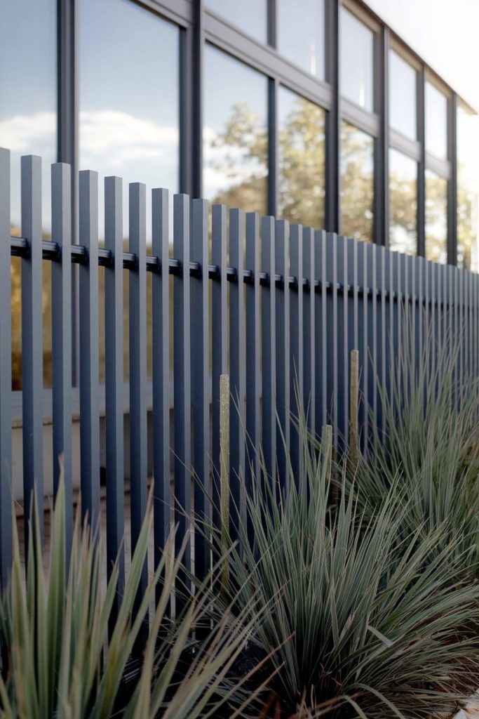 Tall blue fence with vertical slats in front of large reflective windows and green plants.