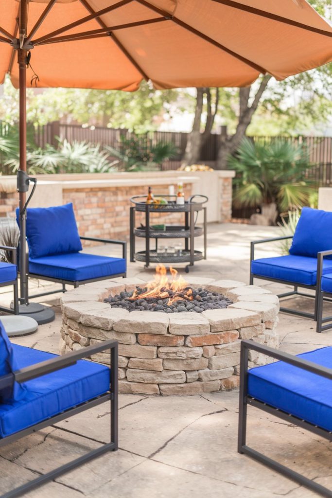 Patio scene with a round stone fire pit and blue upholstered chairs under a parasol surrounded by trees. There is a drinks cart in the background.