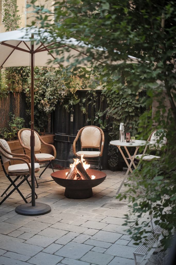 A cozy outdoor patio with a fire pit surrounded by chairs and a table with drinks, shaded by an umbrella and surrounded by foliage.