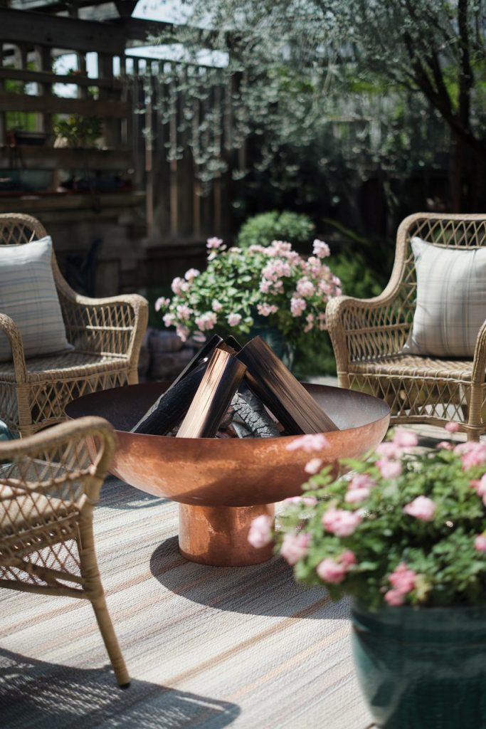 Outdoor seating area with wicker chairs and cushions surrounding a copper fire pit. In the background you can see pink flowers in pots and a wooden structure with hanging plants.