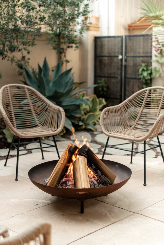 Two wicker chairs sit in front of a metal fire pit with burning logs on a tiled patio surrounded by plants.