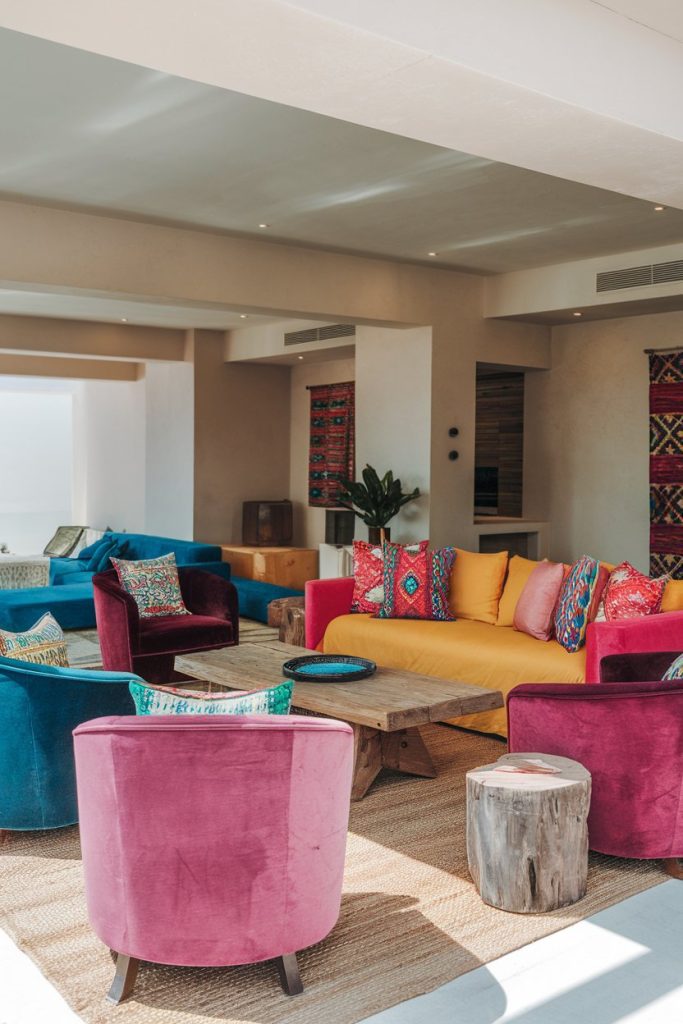 A colorful living room with colorful armchairs, a yellow sofa, patterned pillows, a wooden coffee table and decorative rugs on the walls.