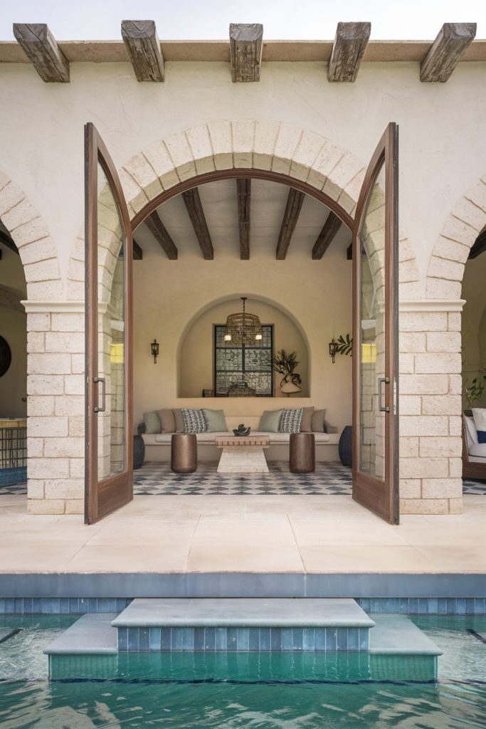 Open arched doors lead to a seating area with a couch and three stools on a tiled floor and overlooking a pool with tiled steps.