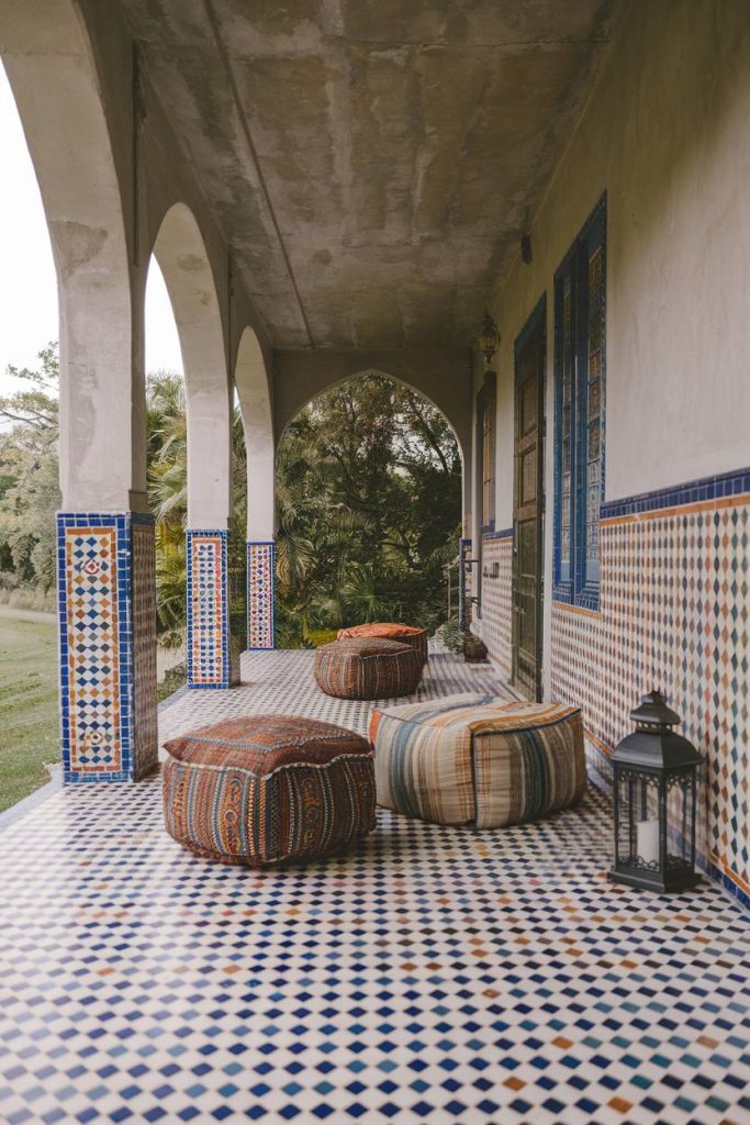 A covered terrace with patterned floor tiles, mosaic columns and three large colorful cushions. There is a lantern on the ground and greenery can be seen in the background.