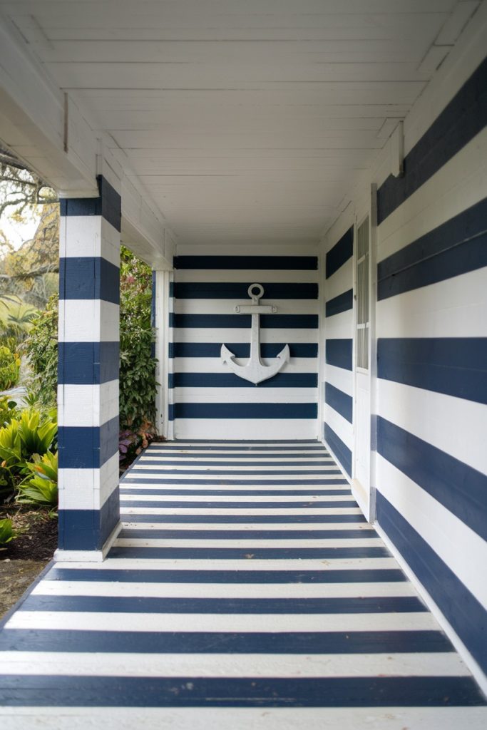 A porch with dark blue and white horizontal stripes on the walls and floor and a white anchor on the back wall. Bushes can be seen outside on the left.