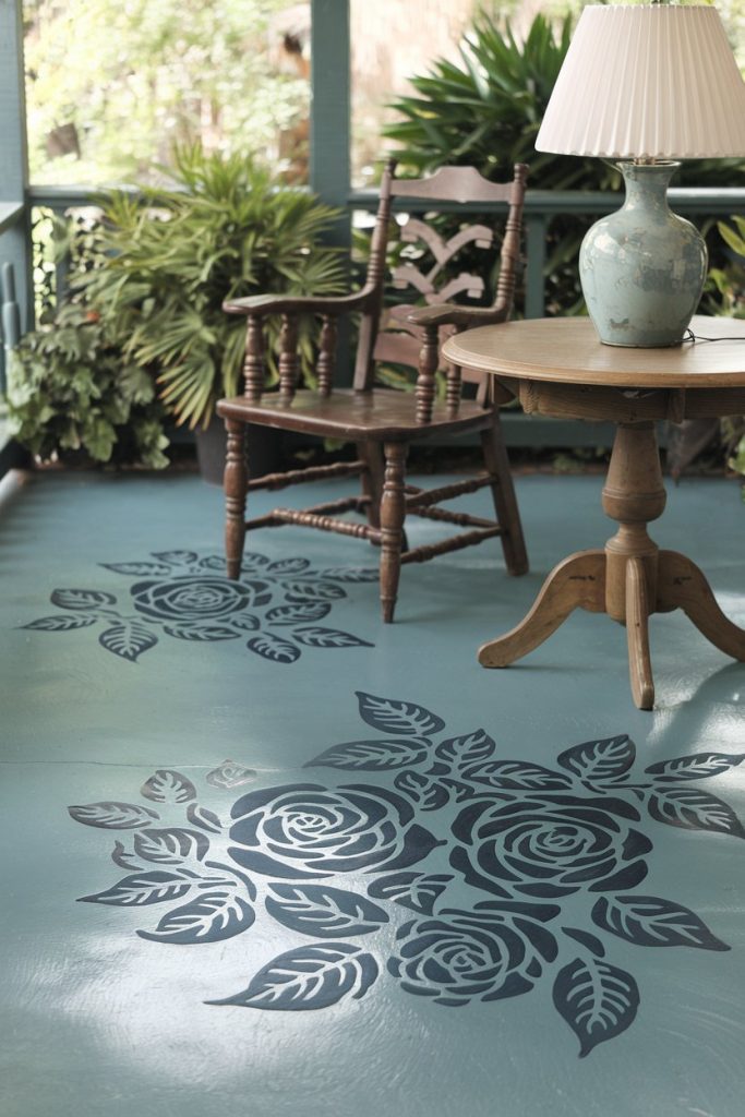 A veranda with a wooden chair, a round table with a lamp and a floor decorated with large floral stencils. Surrounded by various potted plants.