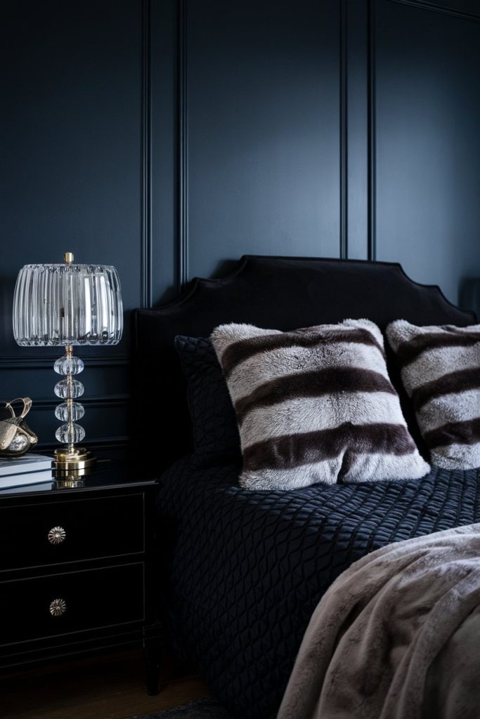 Dark bedroom with a black bed, striped pillows, a textured bedspread and a side table. A lamp made of clear glass with a gold base stands on the table in front of a dark paneled wall.