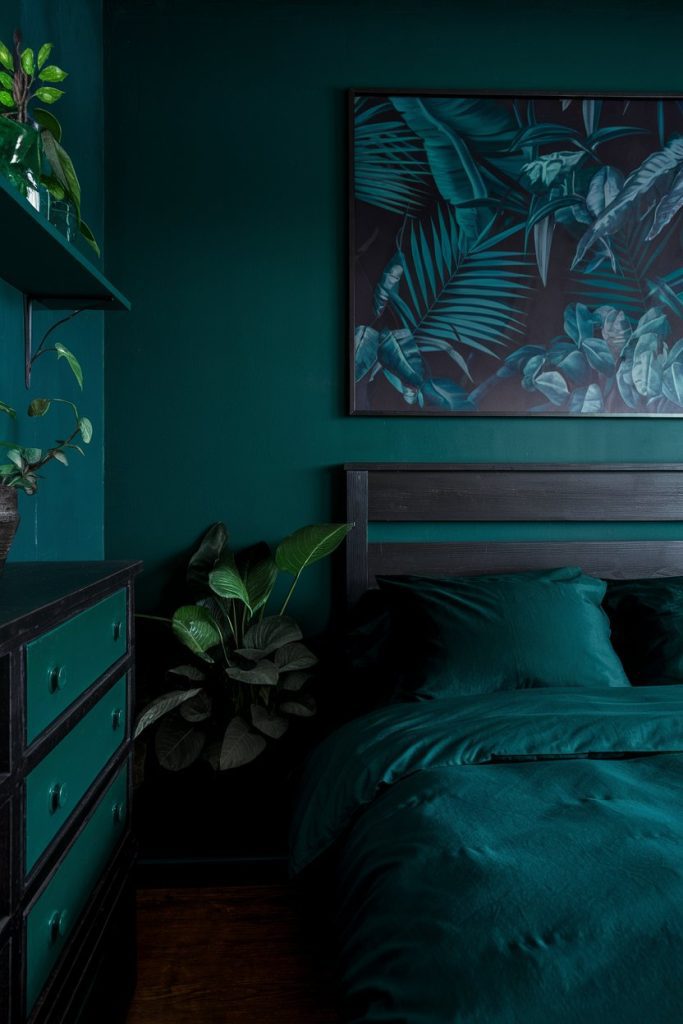 A dark green bedroom with a green bedspread, a dresser, plants and a leaf-themed artwork on the wall.