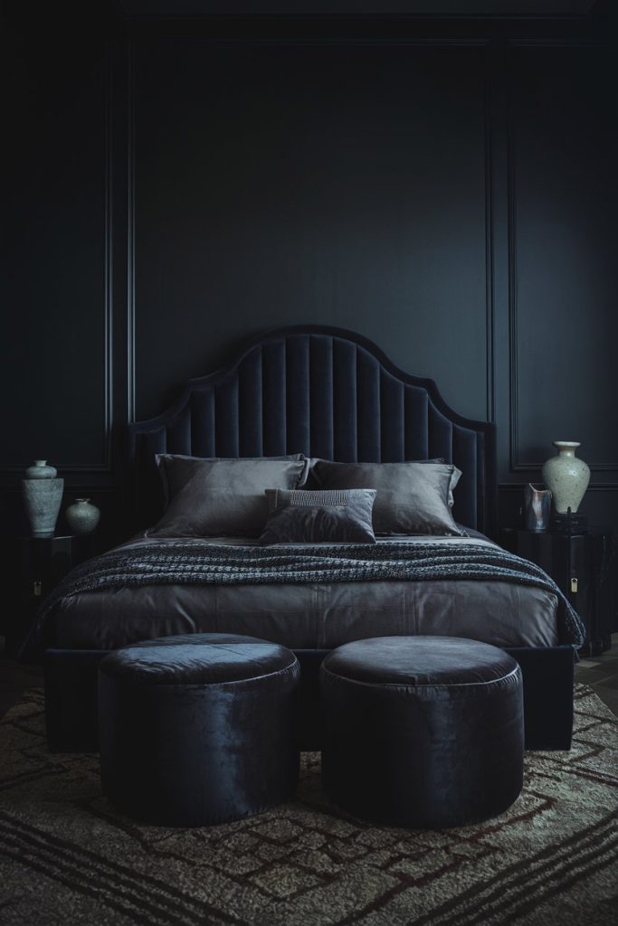 A bedroom decorated in dark tones with a black upholstered bed, matching pillows, two round ottomans at the foot and decorative vases on either side. The walls and bed linen are dark gray.