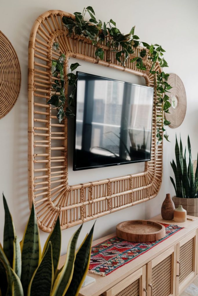 Wall mounted television surrounded by a large wicker rack with hanging plants. Below is a wooden console with a patterned runner, a wooden bowl, pottery and potted plants.