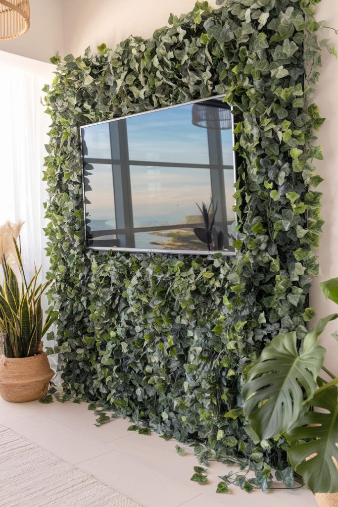 A flat-screen television is mounted on a wall covered in lush green ivy. There are potted plants nearby and a window that offers a scenic view.