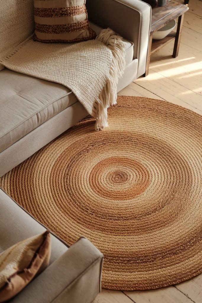 A beige sofa with a textured blanket and cushions, next to a round jute rug on a wooden floor.