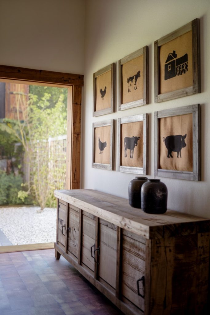 A rustic hallway features a wooden sideboard, two vases and six framed animal silhouette artworks on the wall. A large window opens onto a garden.