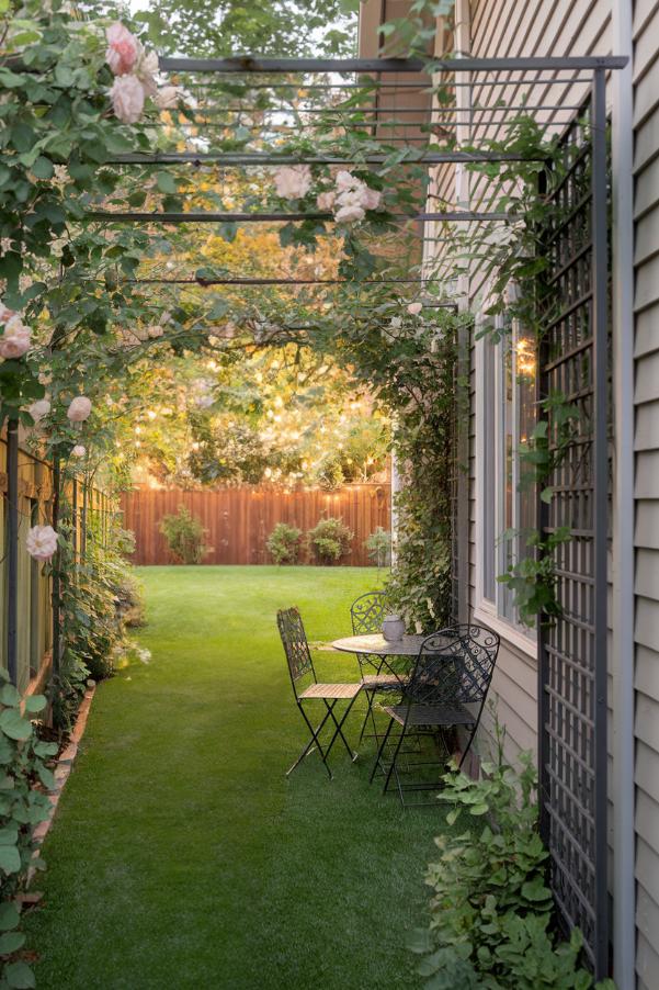 A metal arbor covered with climbing roses arches over a narrow garden path. A small black metal table and two chairs sit next to a house facade that leads to a lawn and a wooden fence.